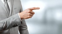 Businessman in light gray suit pointing finger to the side against blurred office background, demonstrating direction or choice in corporate setting.