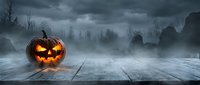 Spooky Halloween jack-o-lantern pumpkin glowing on wooden deck with dark cemetery graveyard background and dramatic stormy sky for seasonal horror decorations.