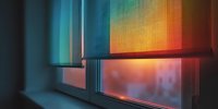 Colorful window blinds with vibrant gradient from blue to orange, casting warm light into room at sunset while partially obscuring urban view outside.
