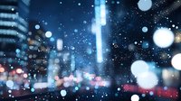 Blurred city lights and falling snow create magical winter atmosphere with bokeh effect in urban night scene, defocused buildings and street illumination in background.