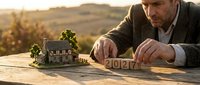 Middle-aged man with light skin tone and dark hair, wearing casual brown clothing, arranging wooden blocks with numbers outdoors at sunset. Real estate planning concept for property investment, home development, and future housing goals.