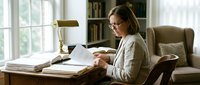 Middle-aged Caucasian woman with glasses working on laptop at wooden desk in bright home office with bookshelf and comfortable chair for remote work productivity.