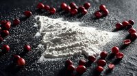 Fresh pomegranate seeds surrounding white flour or powdered sugar on dark textured surface, creating striking color contrast for food photography.