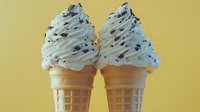 Soft serve vanilla ice cream cones with chocolate cookie crumbles against yellow background, creamy swirled texture and crispy waffle cones for summer dessert menu.