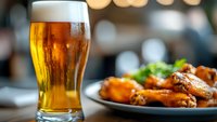 Cold glass of light beer with condensation drops alongside spicy buffalo chicken wings served on dark plate, restaurant setting with bokeh background.