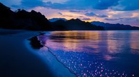 Tranquil beach at twilight with glowing bioluminescent plankton washing ashore, dramatic mountain silhouettes against purple-orange sunset sky reflecting on calm water surface.