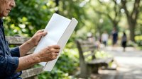 Senior man with light skin tone reading an open book outdoors in a park setting with blurred green trees and pathway in background, suitable for education, leisure, and lifestyle content.