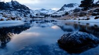 Mountain lake with snow-capped peaks reflecting in calm water during twilight hour. Serene alpine landscape with rocky shoreline and peaceful atmosphere.