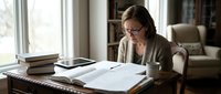 Middle-aged Caucasian woman with glasses reading documents at wooden desk near window in cozy home office with books and natural lighting for study work.
