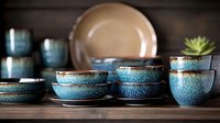 Artisanal ceramic tableware set in turquoise blue glaze with speckled texture displayed on wooden surface, featuring bowls, cups and plates in rustic style.