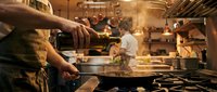 Professional chef cooking in busy commercial kitchen with flames and steam during dinner service preparation.