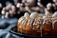 Freshly baked bundt cake with white glaze drizzle and pearl sugar decorations on dark plate, selective focus with blurred cherry blossoms background.