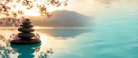 Zen stone stack balanced on calm water surface with cherry blossom branches and mountain landscape at sunset creating peaceful meditation atmosphere.