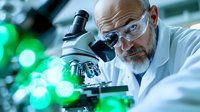 Middle-aged Caucasian male scientist with gray beard wearing protective eyewear and lab coat examines specimen through microscope in modern laboratory with green bokeh lights.