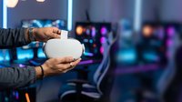 Person holding white smart speaker device in gaming room with colorful neon lighting and blurred gaming chairs in background. Tech gadget display.