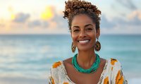 Young African American woman with curly hair smiling at beach sunset, wearing colorful dress and turquoise necklace with gold earrings.