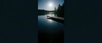 Wooden dock extending into calm lake at dawn with bright sun reflecting on still water and evergreen forest silhouette in background.