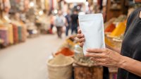 Unrecognizable person holding blank white pouch in spice market with colorful goods and blurred shoppers in background for product packaging mockup.