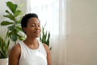 Peaceful African American woman meditating with closed eyes in bright modern room with green plants, practicing mindfulness and wellness at home.