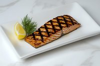 Grilled salmon fillet with distinctive char marks served on white plate, garnished with fresh dill and lemon wedge against marble background.