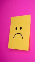 Yellow sticky note with sad face drawn on it against bright pink background expressing disappointment and negative emotions for mental health concepts.