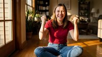 Young Asian woman celebrating success with raised fists sitting cross-legged on wooden floor in modern home interior with natural sunlight streaming through windows.