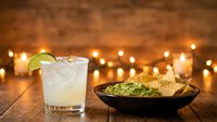 Margarita cocktail with lime slice and salt rim served with tortilla chips and guacamole dip against warm bokeh lights on wooden surface for bar, restaurant, or entertaining.