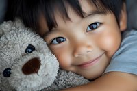 Young Asian child with gentle smile cuddling soft plush teddy bear toy in close-up portrait showing innocence and tender childhood moments.