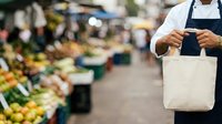 Male vendor of South Asian descent wearing navy apron holding blank canvas tote bag at outdoor farmers market with fresh produce displays in background.