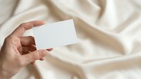 Hand holding blank white business card against soft beige fabric background. Suitable for branding, corporate identity, stationery mockups, and business presentations.