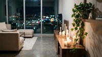 Modern luxury apartment interior with city lights view through floor-to-ceiling windows, featuring minimalist furniture, candles and green plants for cozy ambiance.