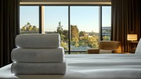 Fresh white folded towels stacked on hotel bed with panoramic window view, morning sunlight streaming in, creating luxurious hospitality atmosphere.