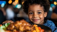 Young African American boy smiling at dinner table with plate of roasted chicken and vegetables, warm bokeh background creates cozy restaurant atmosphere.