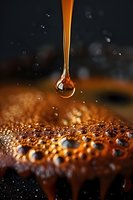 Golden liquid drop falling onto textured surface with water droplets creating dramatic macro photography effect against dark background with bokeh highlights.