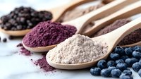 Colorful superfood powders in wooden spoons with fresh blueberries and black peppercorns on light background, healthy organic supplements for smoothies.
