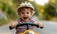Joyful toddler boy wearing safety helmet laughs while riding bicycle on summer day, dressed in red striped t-shirt, capturing pure childhood happiness outdoors.