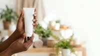 Young woman with dark skin tone holding blank white cosmetic tube for skincare, lotion, or sunscreen product display against blurred botanical background.