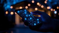 Silhouetted hands holding smartphone with glowing blue screen against blurred bokeh lights background creating magical evening atmosphere.
