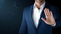 Businessman in navy suit showing stop gesture with raised palm against dark background, conveying boundary setting in professional environment.