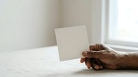 Senior man with dark skin tone resting hand near blank cream card on white marble table by window, suitable for greeting cards, invitations, and stationery mockups.