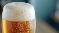 Frothy white foam crowns golden beer in glass with rising bubbles against blurred background, macro shot capturing refreshing carbonation and rich amber color.