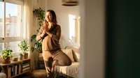 Young woman enjoying morning coffee in cozy sunlit apartment with plants and natural light streaming through windows creating peaceful atmosphere.