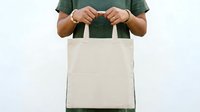 Unrecognizable person with medium skin tone wearing green shirt holding blank natural canvas tote bag for branding mockups and retail presentations.