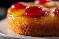 Golden pineapple upside-down cake with glossy caramelized fruit topping and cherries on white plate. Homemade dessert ready for bakery menu or recipe blog.