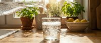 Glass of fresh water pouring into cup on sunny kitchen windowsill with potted plants and fruit bowl, ideal for wellness and healthy lifestyle content.