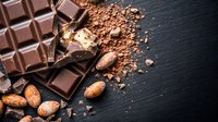 Dark chocolate bar with cocoa powder and roasted cacao beans scattered on black slate background, dramatic lighting creates appetizing mood for food photography.