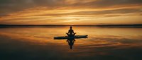 Silhouette of person meditating in lotus position on paddleboard at sunset with golden sky reflection on calm water surface creating peaceful zen moment.