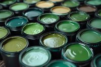 Multiple paint cans filled with various shades of green and olive colors arranged in rows, showing glossy wet surfaces and metallic container edges.