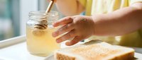 Young woman with medium skin tone reaching for a glass jar of lemonade or iced tea during breakfast, with toast on white table and blurred background.