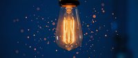 Vintage Edison bulb glowing with warm golden light against deep blue background, surrounded by floating sparkling particles and bokeh effects, symbolizing innovation and creative ideas.